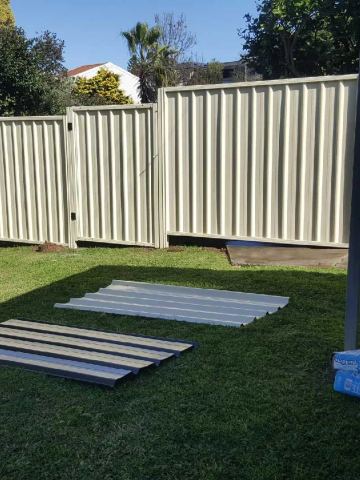 Cream Colorbond privacy fence across a lawn with leftover sheets and a roller door beyond
