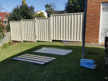Cream Colorbond privacy fence across a lawn with leftover sheets and a roller door beyond