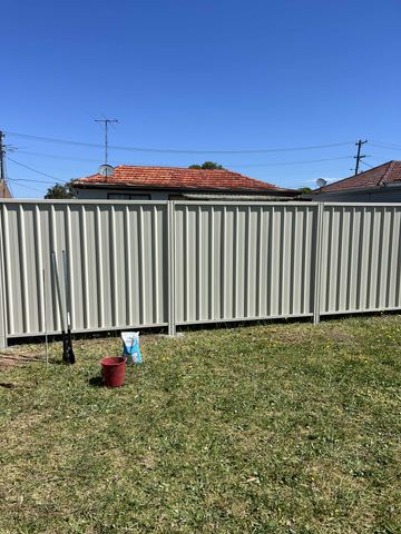 Light grey Colorbond privacy fence across a lawn under a clear sky, with a bucket left on the grass
