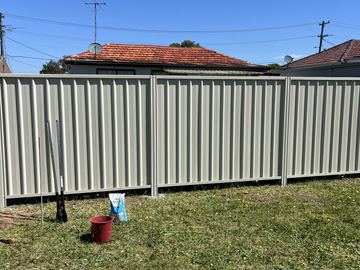 Light grey Colorbond privacy fence across a lawn under a clear sky, with a bucket left on the grass