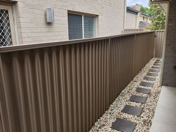Looking along a cream Colorbond privacy fence beside a pebble path and house windows