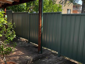 Green Colorbond privacy fence beside a timber pergola post on paving
