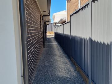 Looking along a dark Colorbond privacy fence beside a pebble path under an eave