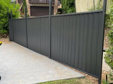 Dark panels on tall black posts follow a grey concrete pad toward a brick house with cream fencing visible beyond.
