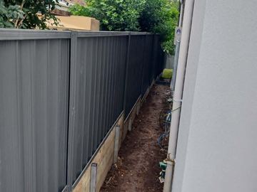 The scene shows a mid grey corrugated steel fence where concrete paving runs alongside.