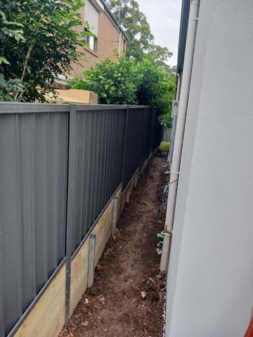 The scene shows a mid grey corrugated steel fence where concrete paving runs alongside.