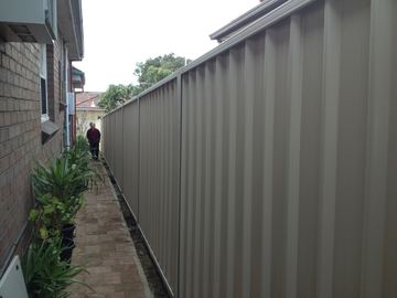 The scene shows a mid grey corrugated steel fence where residential buildings appear beyond the line.
