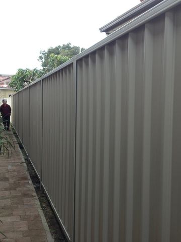 The scene shows a mid grey corrugated steel fence where residential buildings appear beyond the line.