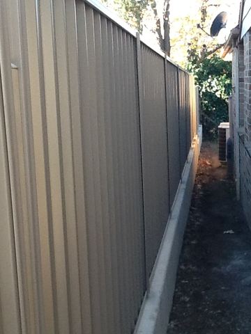 Cream Colorbond privacy fence tight against a brick side wall with a narrow dirt gap