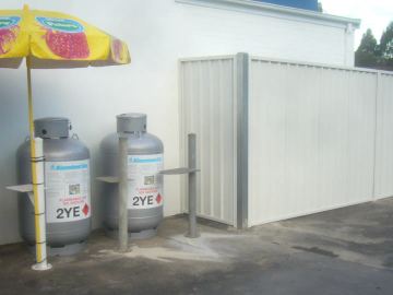 A cream corrugated steel enclosure with a wide gate shields equipment next to Kleenheat gas bottles on an asphalt forecourt.