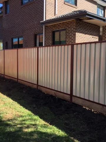 Cream Colorbond privacy fence with brown posts on a timber plinth beside a two-storey brick house