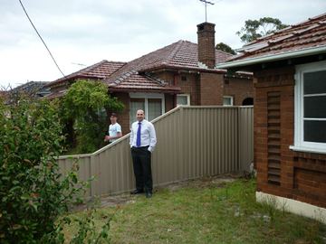 Cream Colorbond privacy fence beside a brick cottage with a person standing on the lawn