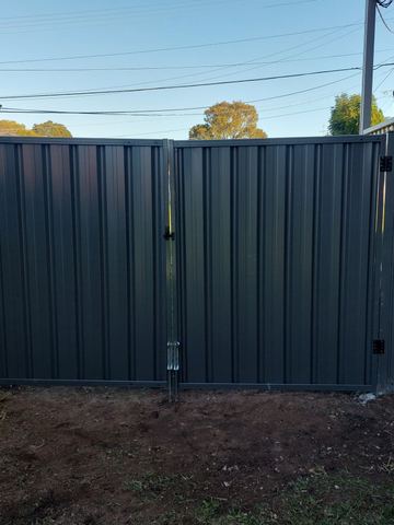 Charcoal double gates with drop bolts stand on fresh soil beside a red brick wall and overhead wires.