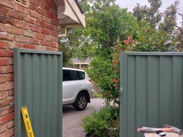 Pale-green corrugated panels with posts flank a brick house driveway where a white SUV is parked and tools sit on a stool.