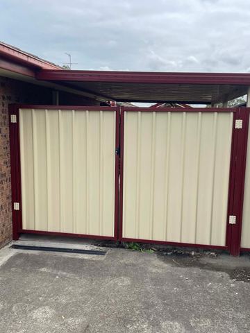 Cream corrugated double gates in heritage-red frames open onto a concrete drive beside a brick carport.