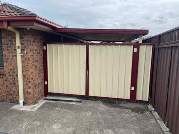 Cream corrugated double gates in heritage-red frames open onto a concrete drive beside a brick carport.