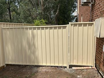 Cream gate and stepped panels on sleepers run along bare dirt beside a brick wall and gum trees.