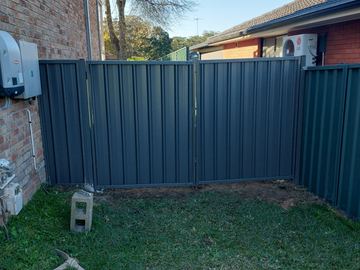 Wide charcoal double gates span between brick walls with solar gear on one side and an air conditioner on the other.