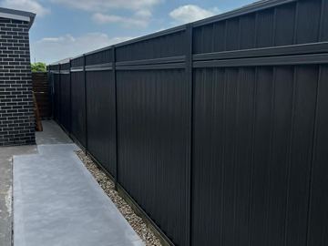 Charcoal panels with a top rail border a concrete walkway separated by pebbles from a dark brick house.