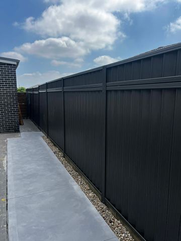 Charcoal panels with a top rail border a concrete walkway separated by pebbles from a dark brick house.