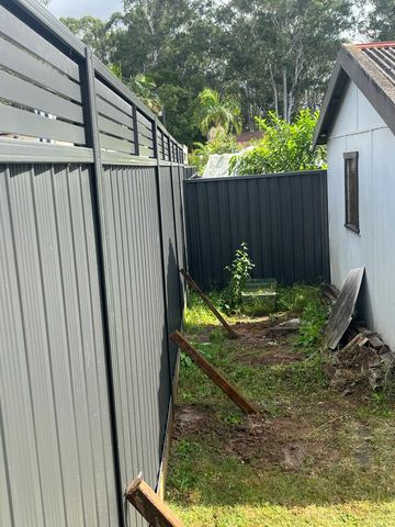 Charcoal panels with slat toppers and propped stakes stand beside a white garden shed on patchy side-yard soil.