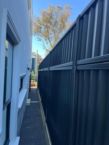 Charcoal tall panels on timber sleepers line a dark paved side passage beside a rendered house and window.