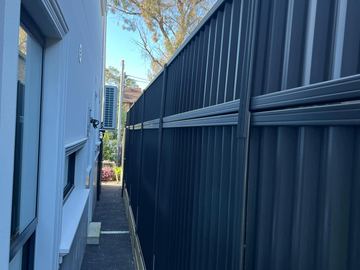 Charcoal tall panels on timber sleepers line a dark paved side passage beside a rendered house and window.