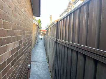 Dark bronze corrugated panels with a mid-rail line a textured concrete side path between brick houses.
