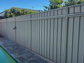 Cream panels and a latched gate run beside travertine pool coping and white pebbles with trees overhead.