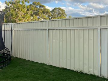 Cream panels with a narrow top band line a lawn with a trampoline edge and cloudy sky above trees.
