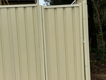 Cream gate and panels enclose a dirt side access with a brick house and trees beyond the latch.