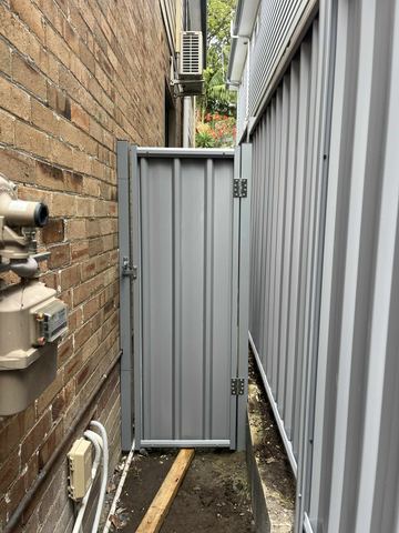 Silver-grey gate and fence fill a tight alley between tan brick walls with gas meter pipes and palm foliage above.