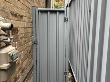 Silver-grey gate and fence fill a tight alley between tan brick walls with gas meter pipes and palm foliage above.