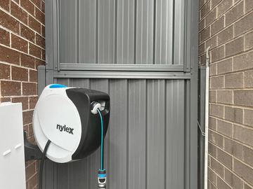 Charcoal gate with upper infill panel sits between brick walls near a wall-mounted hose reel and tap.