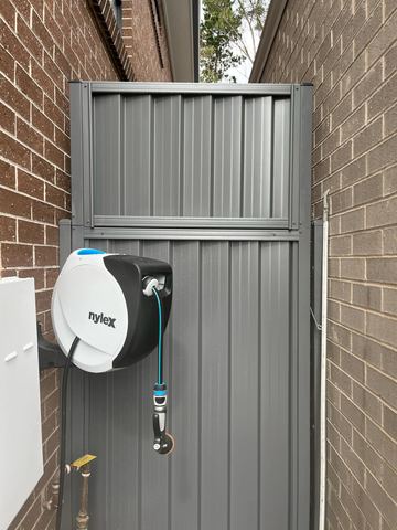 Charcoal gate with upper infill panel sits between brick walls near a wall-mounted hose reel and tap.