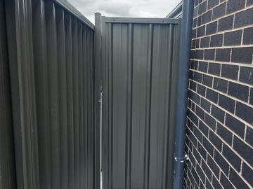Charcoal gate closes a side path beside dark brick and a downpipe on grey concrete under cloudy skies.
