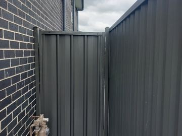 Charcoal gate and tall panel attach to dark brick beside a gas meter on a paved side access.