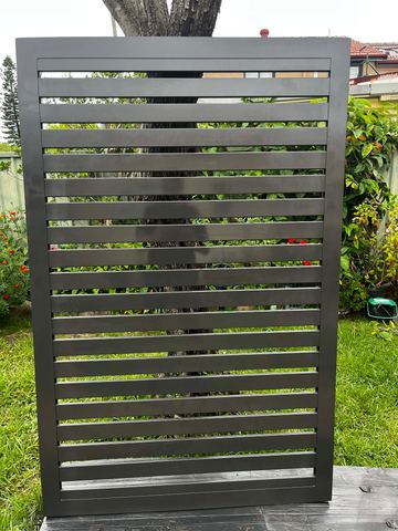 A freestanding charcoal aluminium slat panel stands on grass in a garden bed backed by trees and roof tiles.