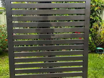 A freestanding charcoal aluminium slat panel stands on grass in a garden bed backed by trees and roof tiles.