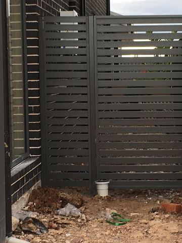 Charcoal slat gate and fixed panel bolt to dark brick on rocky soil with an electrical box behind.