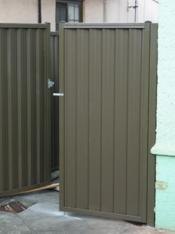 The scene shows a mid grey corrugated steel fence where a gate sits in the run, concrete paving runs alongside.