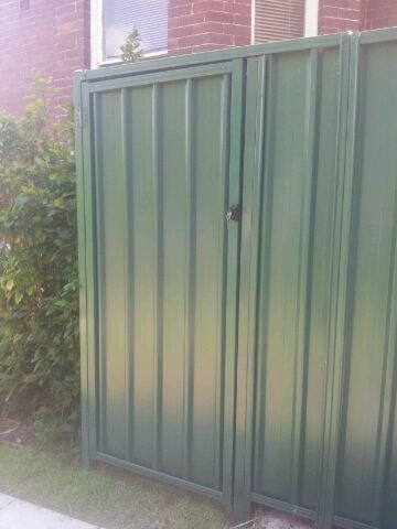 The scene shows a mid grey corrugated steel fence where a gate sits in the run, concrete paving runs alongside.