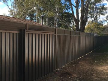 Deep bronze panels and a matching gate carry a continuous louvre extension along a dirt garden bed with gum trees.