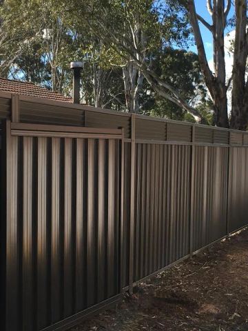 Deep bronze panels and a matching gate carry a continuous louvre extension along a dirt garden bed with gum trees.