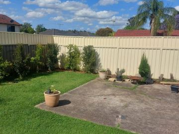 Cream panels enclose a lawn corner with shrubs, a fire pit on pavers and neighbouring roofs beyond.