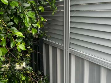 Shale-grey corrugated sheets with a louvre topper meet a leafy hedge beside a black tubular fence corner.