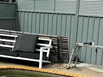 Basalt-grey panels with a louvre top border a pool deck where outdoor seating and a ladder sit on terracotta coping.