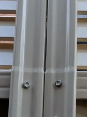 Close-up shows cream panels and louvre extension bolted where two posts meet against brick glimpsed through slats.