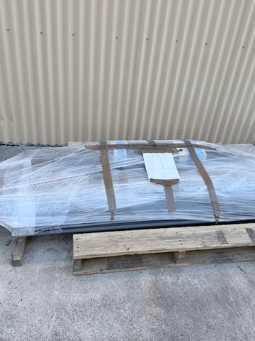 Plastic-wrapped charcoal slat frames sit on a timber pallet against cream corrugated sheeting on a concrete floor.