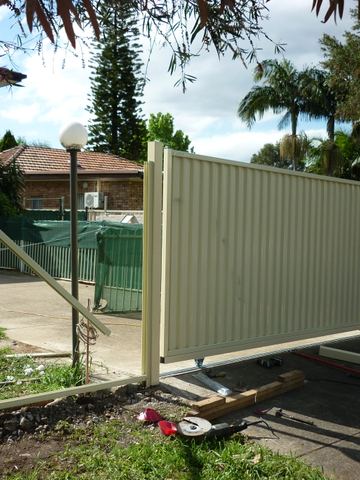 The scene shows a mid grey corrugated steel fence where a gate sits in the run, sleepers form the base.